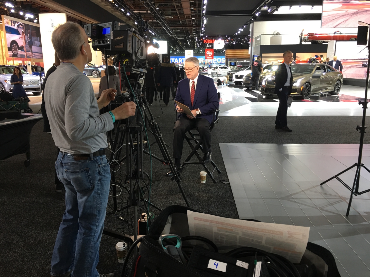 A man is speaking in front of a camera next to vehicles on display at the 2019 Detroit Auto Show