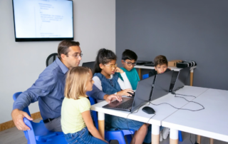 teacher guiding children at laptops with wall monitor