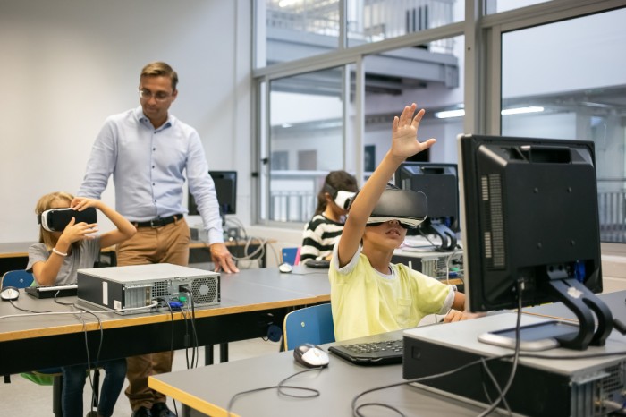 children in computer lab wearing VR headsets beside monitors