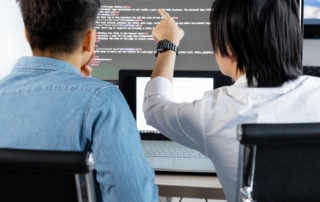 Two people at a desk with a laptop and a large monitor showing code
