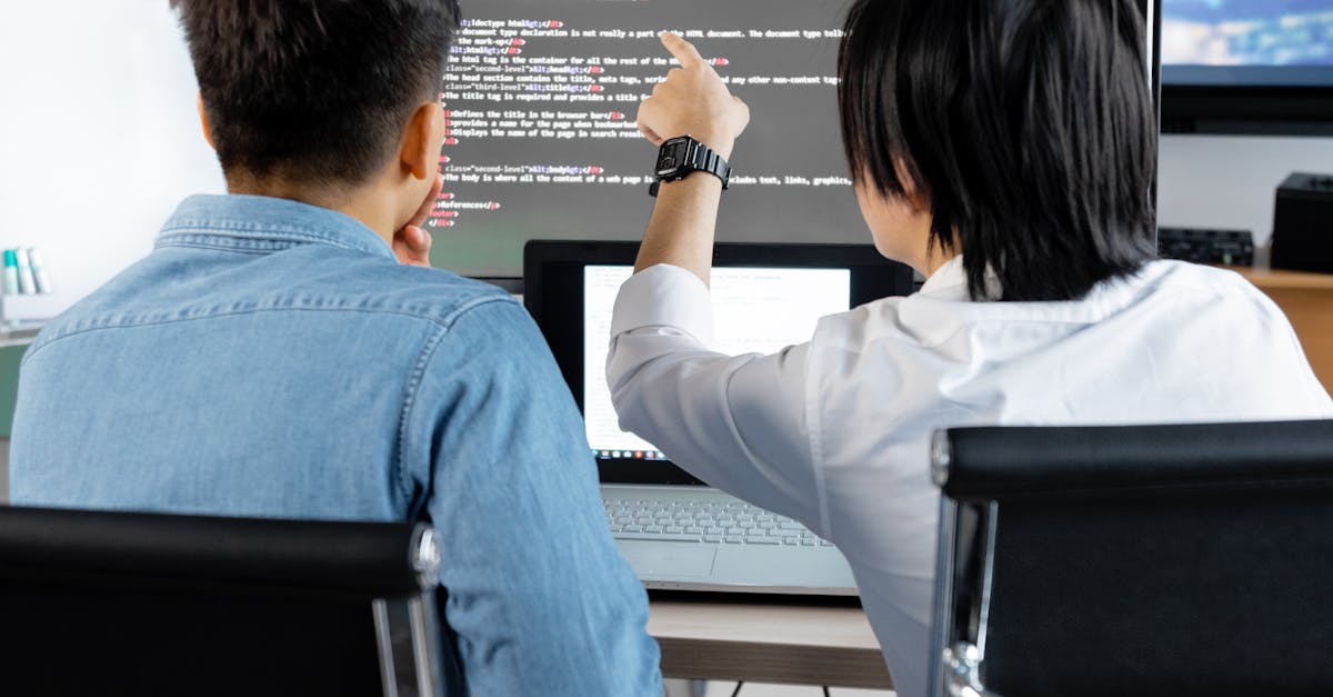 Two people at a desk with a laptop and a large monitor showing code