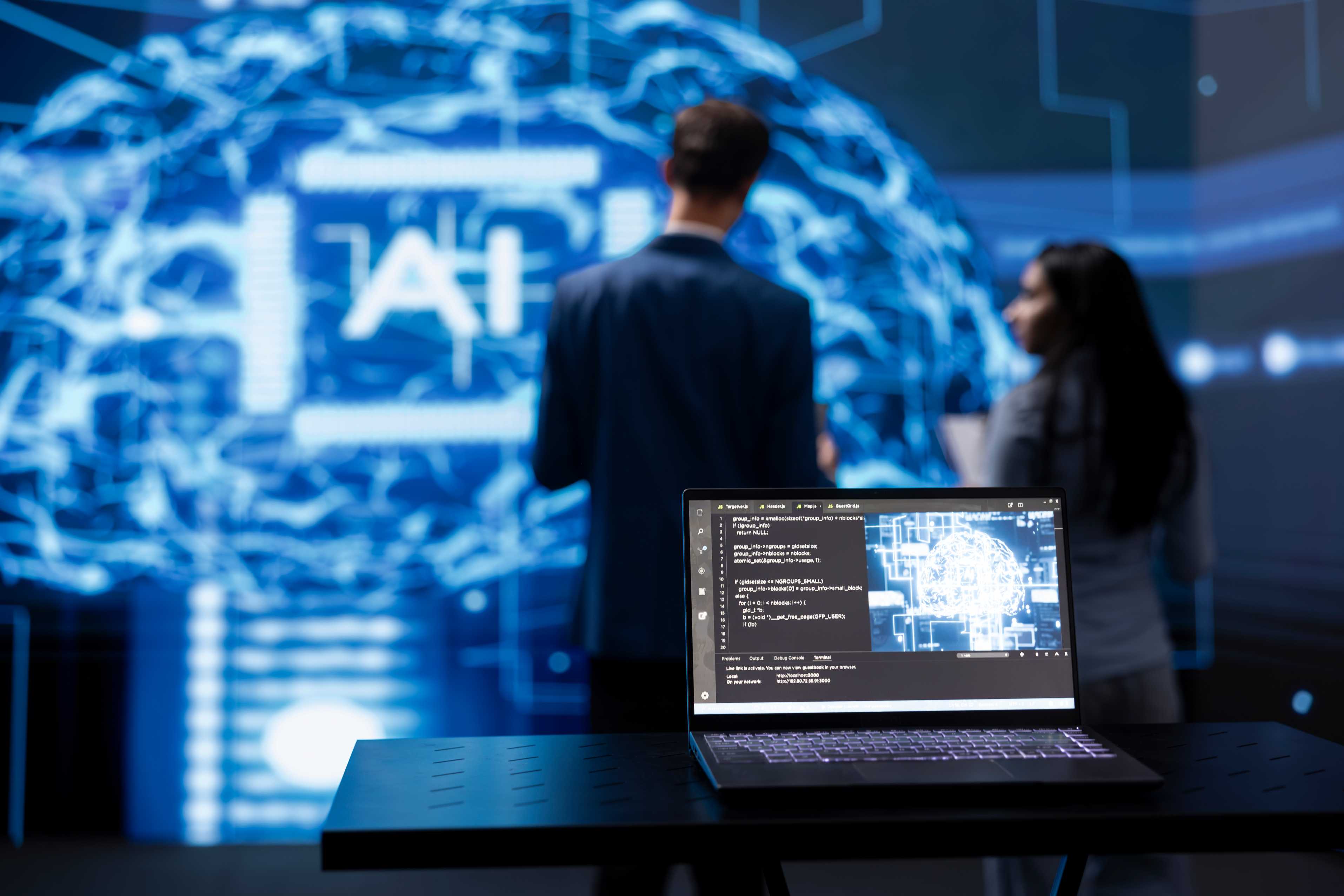 laptop displaying code on table with large screen in background
