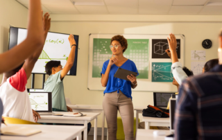 Teacher holding tablet with students raising hands and laptops on desks