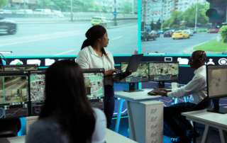 A woman wearing a headset pointing at a bank of wall‑mounted monitors showing maps