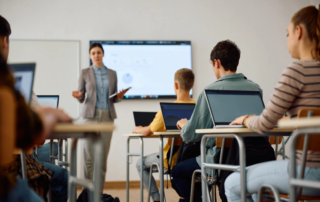 Classroom of students using laptops with teacher presenting on monitor