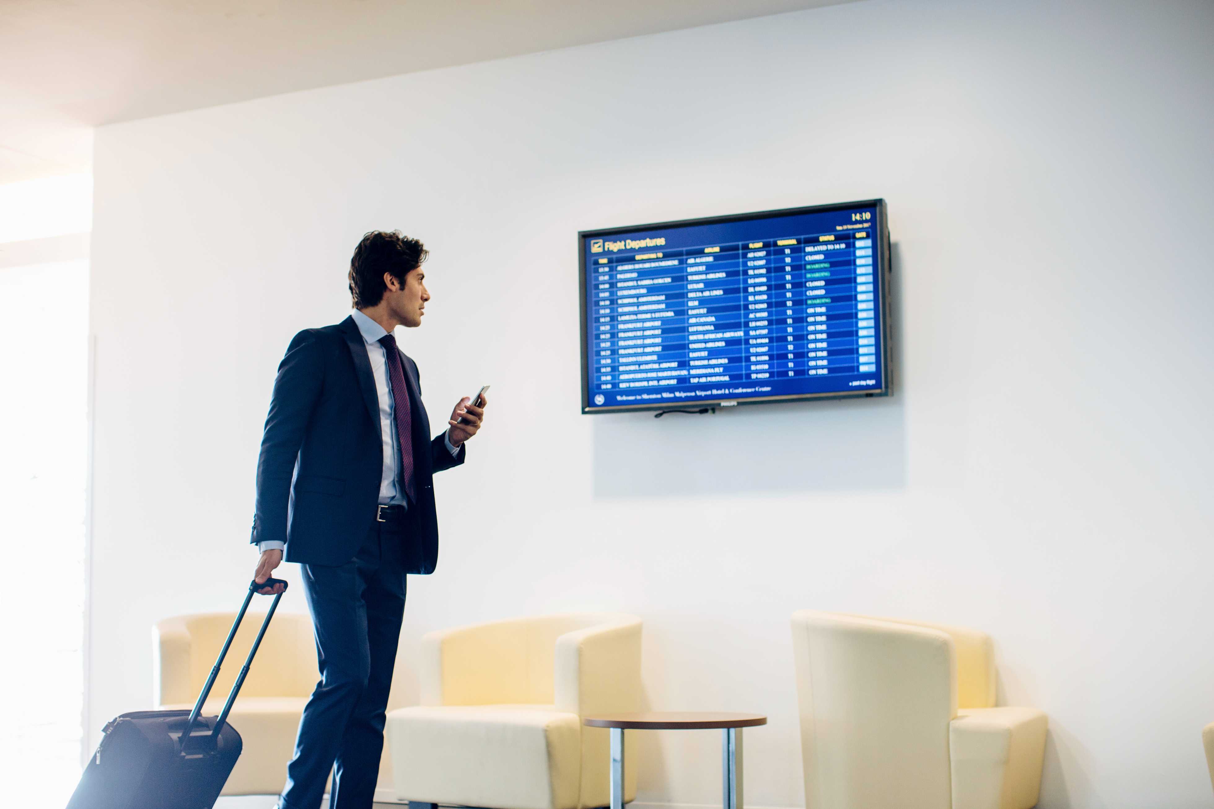 business traveler rolling suitcase checking flight departure screen holding phone near lounge chairs
