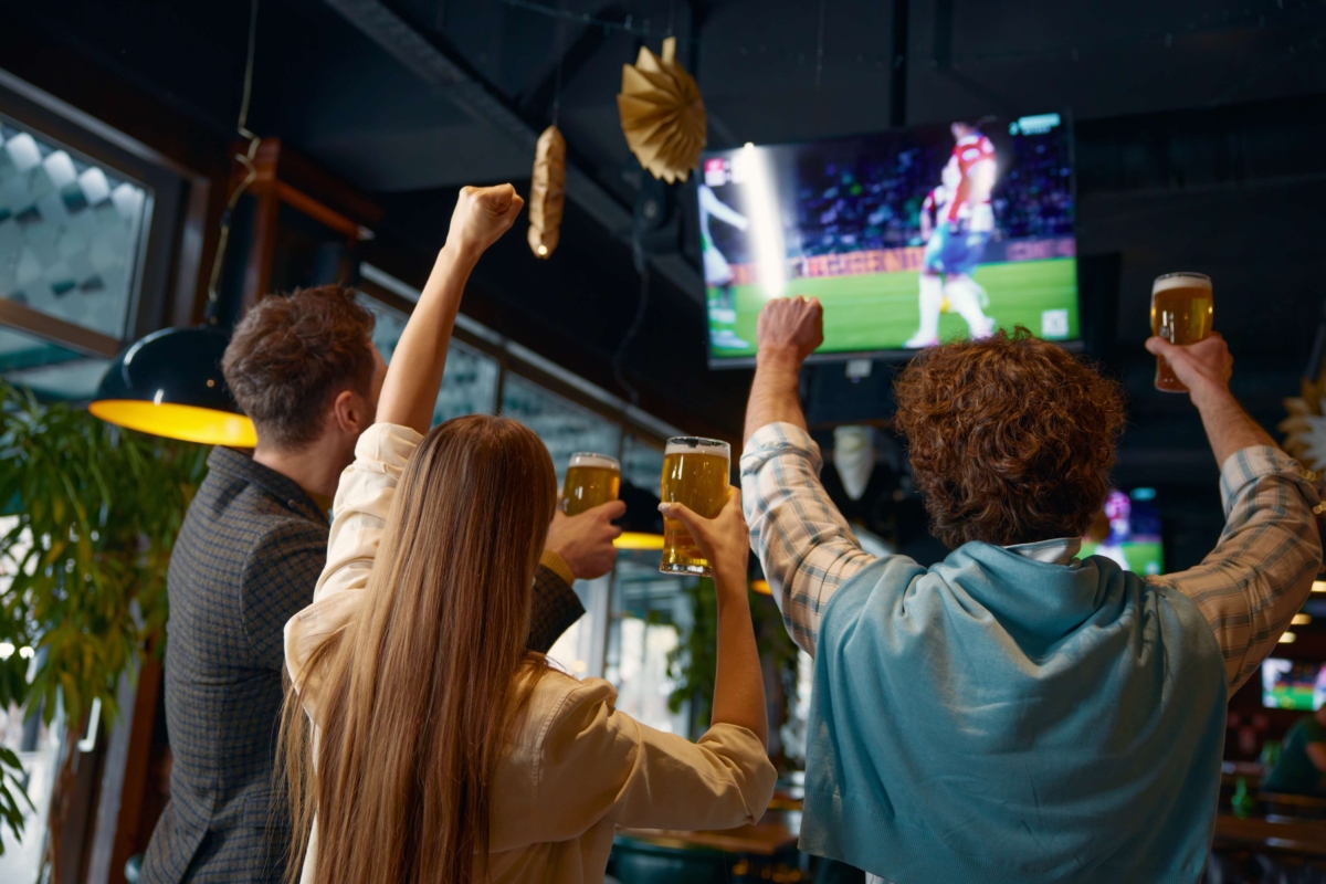 happy-excited-group-of-friends-watching-football-m-2024-12-02-08-01-27-utc (1) group cheering with raised beer glasses watching wall mounted display screen showing soccer match