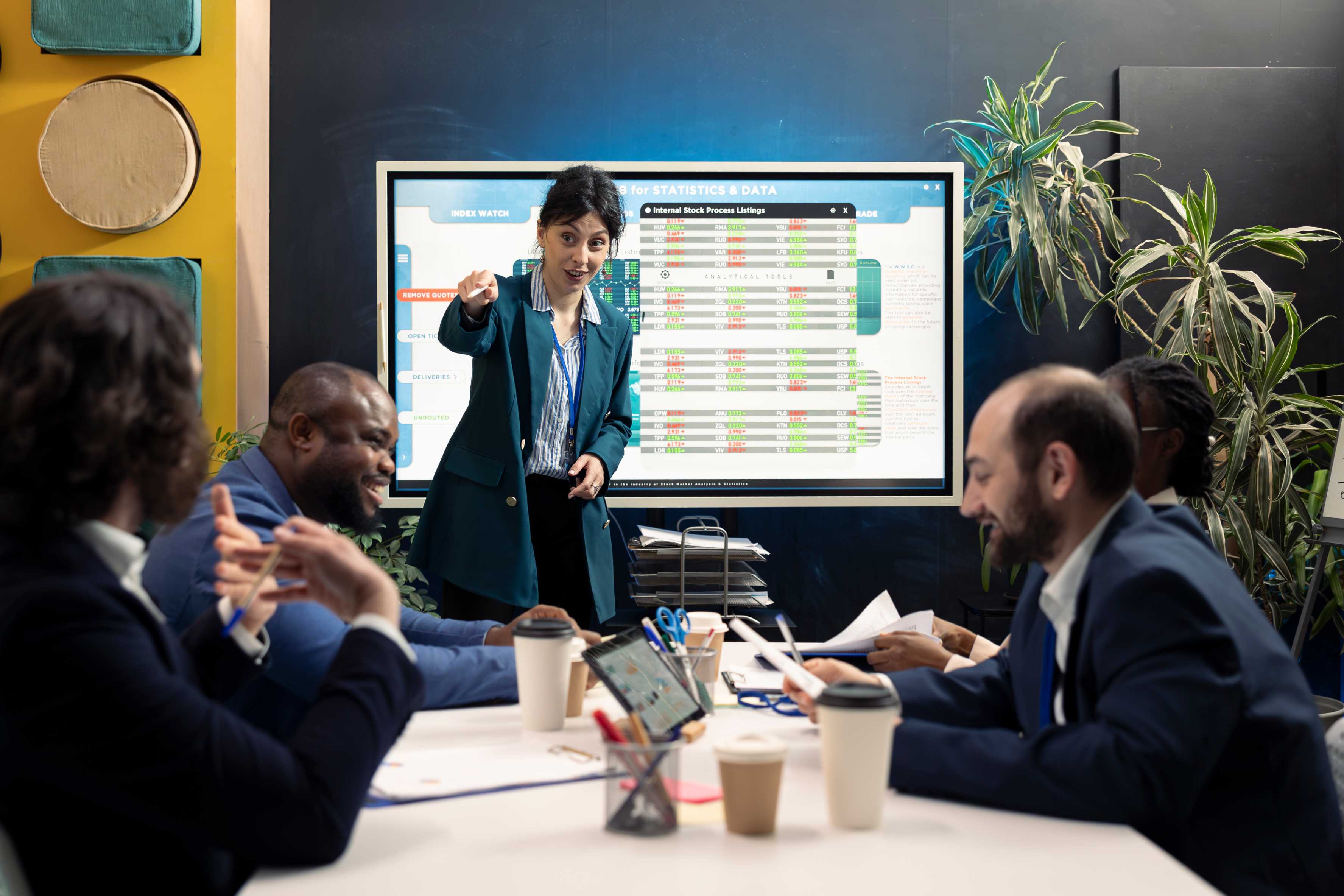 Presenter pointing at large touchscreen display with stock data participants at table with laptops tablets and coffee cups